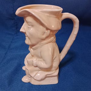 Puede incluir: Jarra de cerámica color crema con forma de hombre sentado con sombrero. El asa es curva y el pico está en la parte superior del sombrero. La jarra tiene un diseño vintage detallado.