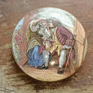 May include: A round, antique ceramic plate featuring a colorful illustration of two figures in period clothing. The man, in a burgundy coat, whispers to the woman. The plate is trimmed in gold and has the text "UNCLE TOBY" at the bottom.