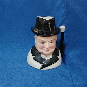 Puede incluir: Una taza de cerámica con forma de cabeza de hombre, que lleva un sombrero negro y un traje negro. La taza tiene un asa y está pintada con rasgos faciales realistas, incluyendo mejillas rosadas y ojos azules. La taza está sobre un fondo azul.