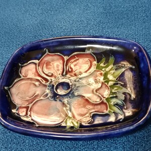 May include: A small, blue ceramic dish with a raised, pink flower design. The flower has a white center and green leaves.