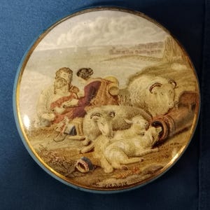 Puede incluir: Una caja de baratijas redonda y antigua con una escena pastoral pintada. La tapa presenta una imagen de personas con ovejas, un barril y un pueblo distante. La caja tiene un borde azul claro con ribete dorado y la palabra "PEACE" en la parte inferior.