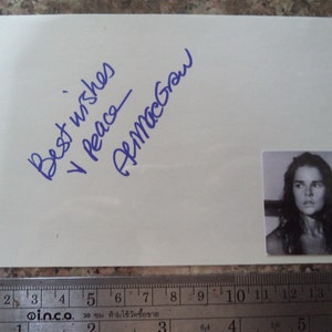 以下が含まれることがあります： 青いインクで「最高の願いと平和」と手書きされた白いカードと、「アリ・マックグロー」という署名。右下にアリ・マックグローの小さな白黒写真があります。