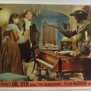 Könnte beinhalten: Ein Vintage-Filmstill aus "Dr. Syn, alias The Scarecrow" mit Schauspielern in historischen Kostümen. Die Szene zeigt ein Cembalo, eine Vogelscheuchen-ähnliche Figur und ein Paar in einem Raum mit Kronleuchter. Der Text unten lautet "Walt Disney's Dr. Syn alias 'The Scarecrow' Patrick McGoohan Technicolor."