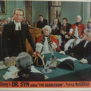 May include: A black and white movie still from the film "Dr. Syn" starring Patrick McGoohan. The scene takes place in a courtroom with a group of men in period clothing. The main character, Dr. Syn, is standing in the center of the image, addressing the court.