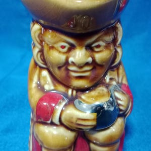 May include: A ceramic figurine of a smiling man with red eyes, wearing a hat and holding a small bowl. The figurine is primarily brown with red and black accents. The object is a small decorative item.