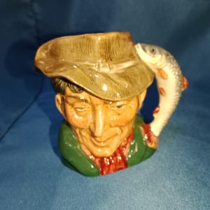 Op de afbeelding: Een keramische mok in de vorm van een man met een groene hoed en jas, met een rode sjaal. Een vis is aan de zijkant van de mok bevestigd als handvat. De mok is beschilderd met realistische details en kleuren.