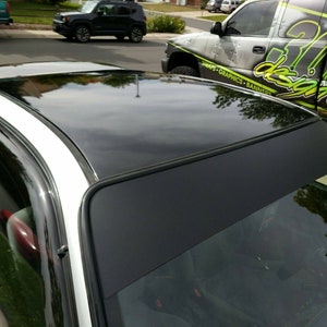 May include: A white car with a black vinyl wrap on the roof and windshield. The wrap has a black border around it. The car is parked in a driveway.