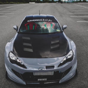 May include: A gray sports car with a black hood and a "loweredlifestyle" windshield banner. The car is parked in an empty parking lot.