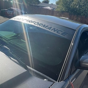May include: A grey car with a windscreen banner displaying "FORD PERFORMANCE" in black text. The car is parked outdoors on a sunny day. Another car is visible in the background.