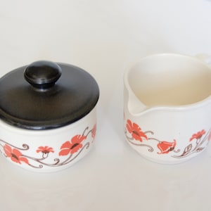 May include: A white ceramic sugar bowl and creamer set with a floral design. The sugar bowl has a black lid. The creamer has a spout and a handle.