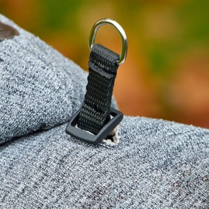 Op de afbeelding: Close-up van een zwarte band met een zilveren D-ring bevestigd aan een grijze stof. De band heeft een zwarte plastic gesp.