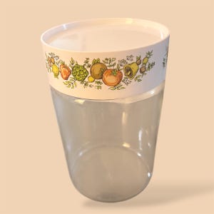 May include: A clear, cylindrical container with a white lid. The container's upper section features a white band adorned with a colorful fruit and vegetable pattern. The design includes illustrations of lemons, tomatoes, and other produce.