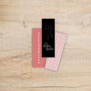 May include: Three rectangular bookmarks on a wooden surface. One is black with a line drawing of a person reading and the text "The darker the better". Another is pink with the text "STFUATTDLAGG". The third is pink with the text "HOT GIRLS READ SMUT".