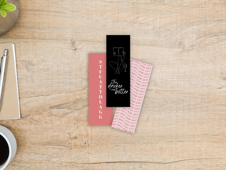May include: Three rectangular bookmarks on a wooden surface. One is black with a line drawing of a person reading and the text "The darker the better". Another is pink with the text "STFUATTDLAGG". The third is pink with the text "HOT GIRLS READ SMUT".