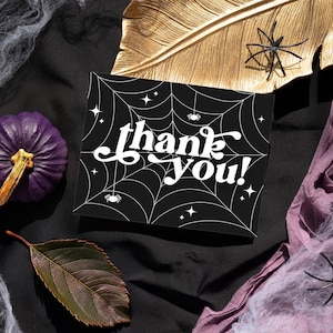 May include: A black card with a white spiderweb design and the text "thank you!" in a retro font. The card is surrounded by purple fabric, gold leaf, and white spiderwebs.