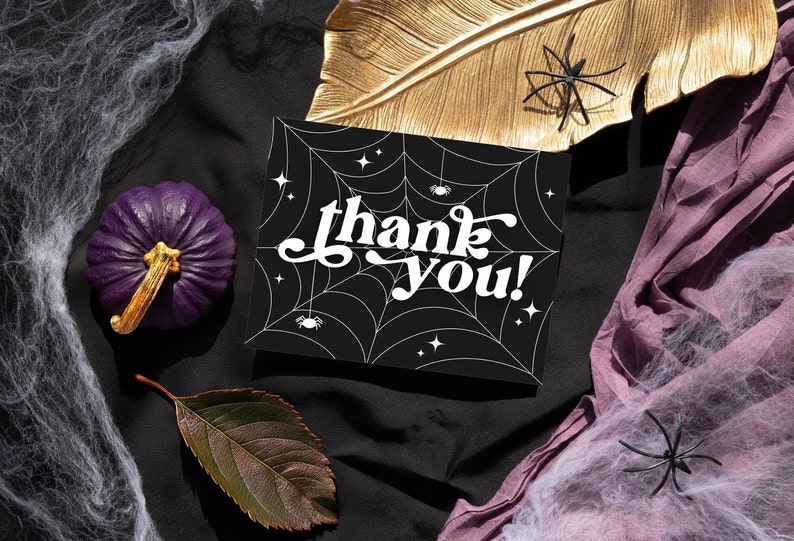 May include: A black card with a white spiderweb design and the text "thank you!" in a retro font. The card is surrounded by purple fabric, gold leaf, and white spiderwebs.