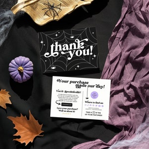 May include: A thank you card with a black and white spiderweb design and the text "thank you!" in white. The card is surrounded by purple fabric, brown leaves, and white spiderwebs.