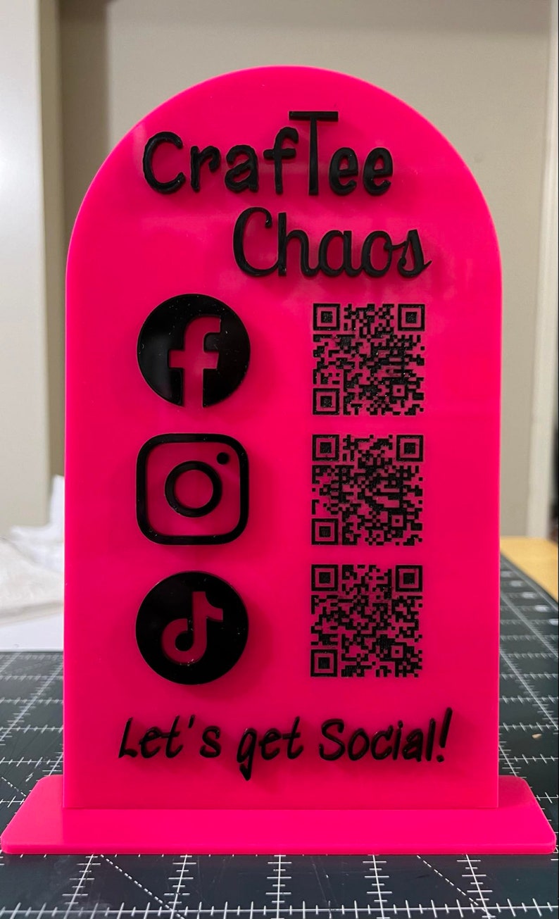 May include: Pink acrylic sign with the text "CraftTee Chaos" and the social media icons for Facebook, Instagram, and TikTok.  The sign also includes three QR codes and the text "Let's get Social!"