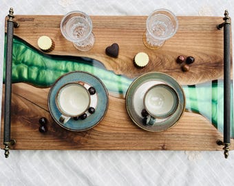 May include: A wooden serving tray with a green and blue epoxy resin inlay. The tray has two brass handles and features two cups and saucers, two shot glasses, and some chocolates.