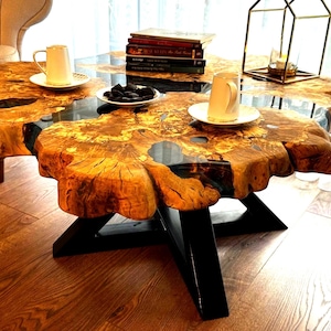 Op de afbeelding: Een ronde houten salontafel met een live edge blad en een zwarte epoxyharsvulling. De tafel heeft een zwart metalen onderstel met een stervormig ontwerp.