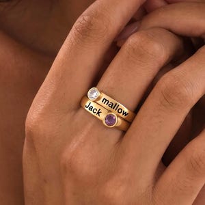 May include: A gold-toned ring featuring two stacked bands. The top band is engraved with the name 'mallow' and set with a clear round stone. The bottom band is engraved with the name 'Jack' and set with a small purple round stone.