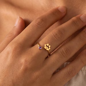 Peut inclure: Une bague ouverte en or dotée d'une petite pierre précieuse ronde violette à une extrémité et d'une breloque en forme de patte découpée avec la lettre H gravée à l'autre extrémité. La bague est en métal doré poli avec un anneau fin et réglable.