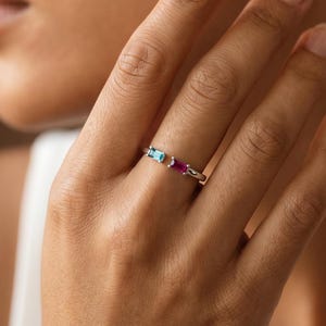 May include: A thin silver-toned ring featuring two small, rectangular-cut gemstones set in a horizontal orientation. One stone is a vibrant light blue, and the other is a deep magenta pink. The band is slender and polished, encircling the finger.