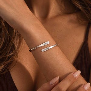 Peut inclure: Un bracelet ouvert de couleur argentée doté de deux extrémités plates et effilées. Chaque extrémité est gravée d'un prénom, Gideon et Raphael, dans une police sans empattement. Le bracelet présente une finition métallique polie.