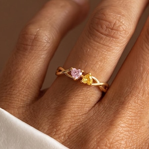 May include: A gold-toned ring featuring a delicate, twisted band design. The ring is adorned with two small, heart-shaped gemstones: one light pink and one yellow, positioned side-by-side at the centre of the band's crossover detail.
