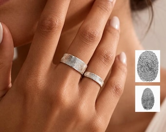 May include: Two silver rings featuring engraved fingerprint patterns on the outer bands. The wider ring is worn on the middle finger, while the thinner ring is worn on the ring finger. Two small square insets show black and white fingerprint designs.