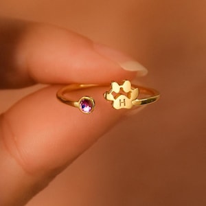 Op de afbeelding: Een goudkleurige ring met een pootafdrukontwerp en een kleine, ronde, paarse edelsteen. De ring is open, waardoor de maat kan worden aangepast. De pootafdruk heeft de letter "H".