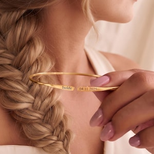 May include: A gold-toned open cuff bracelet featuring a polished finish. The bracelet is engraved with the name Isolde on one end and the date 04.11.2025 on the other end. The metal band has a slender, minimalist profile with a gap in the center.