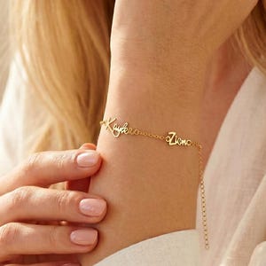 Op de afbeelding: Een goudkleurige metalen kettingarmband met twee afzonderlijke naamhangers in een cursief lettertype. De namen Kaylee en Zion zijn verwerkt in de fijne schakelketting, die aan het uiteinde een verstelbaar verlengstuk bevat.