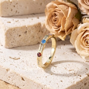 Könnte beinhalten: Ein goldfarbener Ring mit drei kleinen runden Edelsteinen in Blau, Rot und Violett, wobei die Namen Evelyn, Camila und Violet vertikal entlang der Ringschiene eingraviert sind, aufrecht stehend auf einem strukturierten cremeweißen Steinblock.