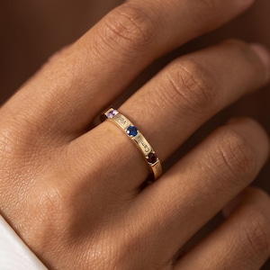Puede incluir: Un anillo de banda en tono dorado con tres pequeñas piedras preciosas redondas y multicolores en violeta claro, azul oscuro y rojo intenso. La superficie metálica está grabada con los nombres Violet y Camila entre las piedras.