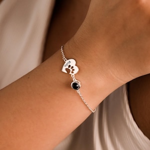 May include: A delicate silver bracelet featuring a paw print charm with the name "Rolo" engraved, and a black gemstone. The bracelet is worn on a wrist.