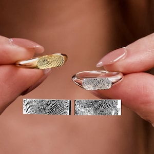 Puede incluir: Dos anillos, uno dorado y otro plateado, cada uno con un diseño de huella dactilar. Los anillos se sostienen en alto, mostrando los detalles únicos de las huellas dactilares. Debajo de los anillos hay dos muestras de huellas dactilares.