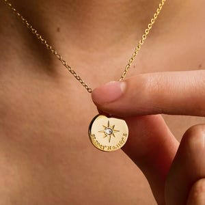 May include: A gold-toned necklace featuring a circular pendant with a central starburst design and a small embedded stone. The pendant is engraved with the text 52.2043 N 0.1167 E, suspended from a delicate gold-toned chain link necklace.
