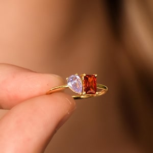 Könnte beinhalten: Ein goldener Ring mit zwei Edelsteinen: einem birnenförmigen Lavendelstein und einem rechteckigen roten Stein. Der Ring wird zwischen den Fingern gehalten und zeigt sein zartes Design und seine leuchtenden Farben. Schmuck.