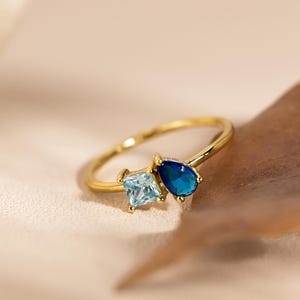 May include: A gold ring with two gemstones: a square, light blue stone and a teardrop-shaped, dark blue stone. The ring is set on a neutral background with a leaf.
