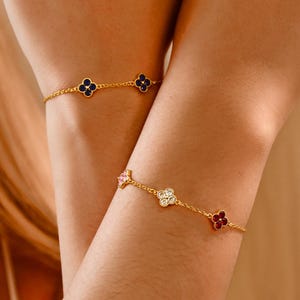 Könnte beinhalten: Zwei goldene Armbänder mit vierblättrigen Kleeblatt-Anhängern. Ein Armband hat blaue Anhänger, das andere hat rosa, weiße und rote Anhänger.