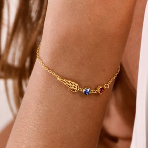 Puede incluir: Una pulsera de cadena dorada con un pequeño dije de ala detallado junto a dos piedras preciosas redondas engastadas, una de color azul vibrante y otra de color rojo intenso, unidas por una delicada cadena de eslabones dorados en la muñeca.