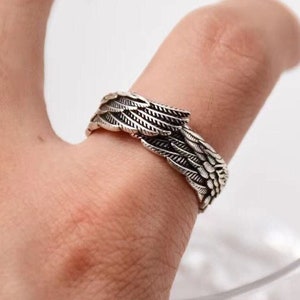 Op de afbeelding: Een zilveren ring met een gedetailleerd vleugelontwerp. De ring wordt aan een vinger gedragen.
