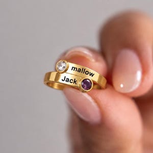 May include: A gold-toned bypass ring featuring two bands, each engraved with a name. One band displays the name "mallow" with a clear round stone, and the other band displays the name "Jack" with a purple round stone set in a bezel.