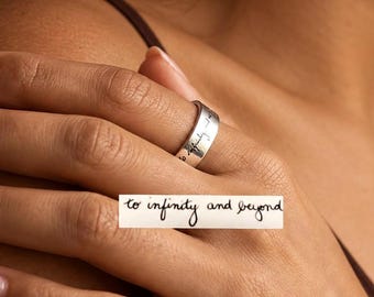 May include: A silver-toned metal band ring featuring the engraved text "to infinity and beyond" in a cursive script. The ring has a smooth, polished finish and a flat profile, showcasing the clear black lettering etched into the metallic surface.