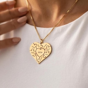Pode incluir: Um colar com um pingente em forma de coração dourado. O pingente é feito de peças de quebra-cabeça com nomes gravados. A palavra "MOM" está no centro. O colar é usado em uma corrente dourada.