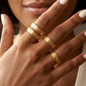 Op de afbeelding: Een hand toont vier gouden ringen, elk gegraveerd met tekst. De ringen zijn van verschillende breedtes en worden op verschillende vingers gedragen. De ringen hebben een gepolijste afwerking en staan tegen een neutrale achtergrond.