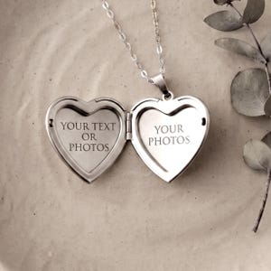 Puede incluir: Un colgante de relicario en forma de corazón de tono plateado en una cadena fina. El relicario está abierto, revelando dos compartimentos interiores. El lado izquierdo está grabado con el texto YOUR TEXT OR PHOTOS y el derecho con YOUR PHOTOS.