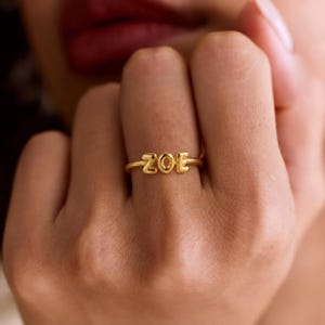 Puede incluir: Un anillo de tono dorado que presenta el nombre ZOE en letras mayúsculas redondeadas y en relieve. La banda es delgada y circular, con las letras posicionadas horizontalmente en la parte frontal del anillo, creando una pieza de joyería personalizada.