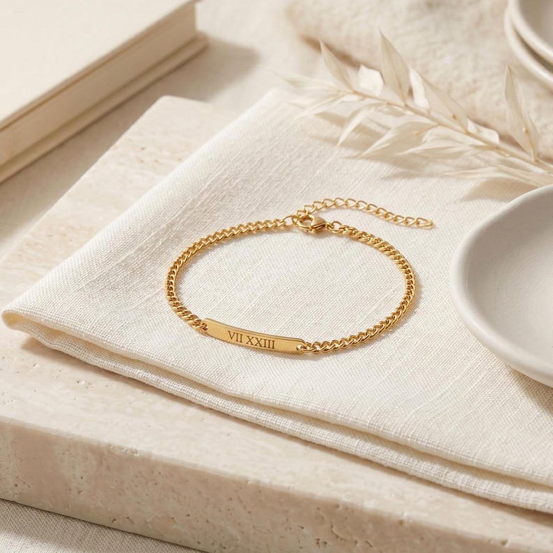 May include: A gold-toned chain bracelet featuring a central flat rectangular bar engraved with the Roman numerals VII XXIII. The bracelet includes a delicate link chain and an adjustable extender section with a small lobster claw clasp closure.